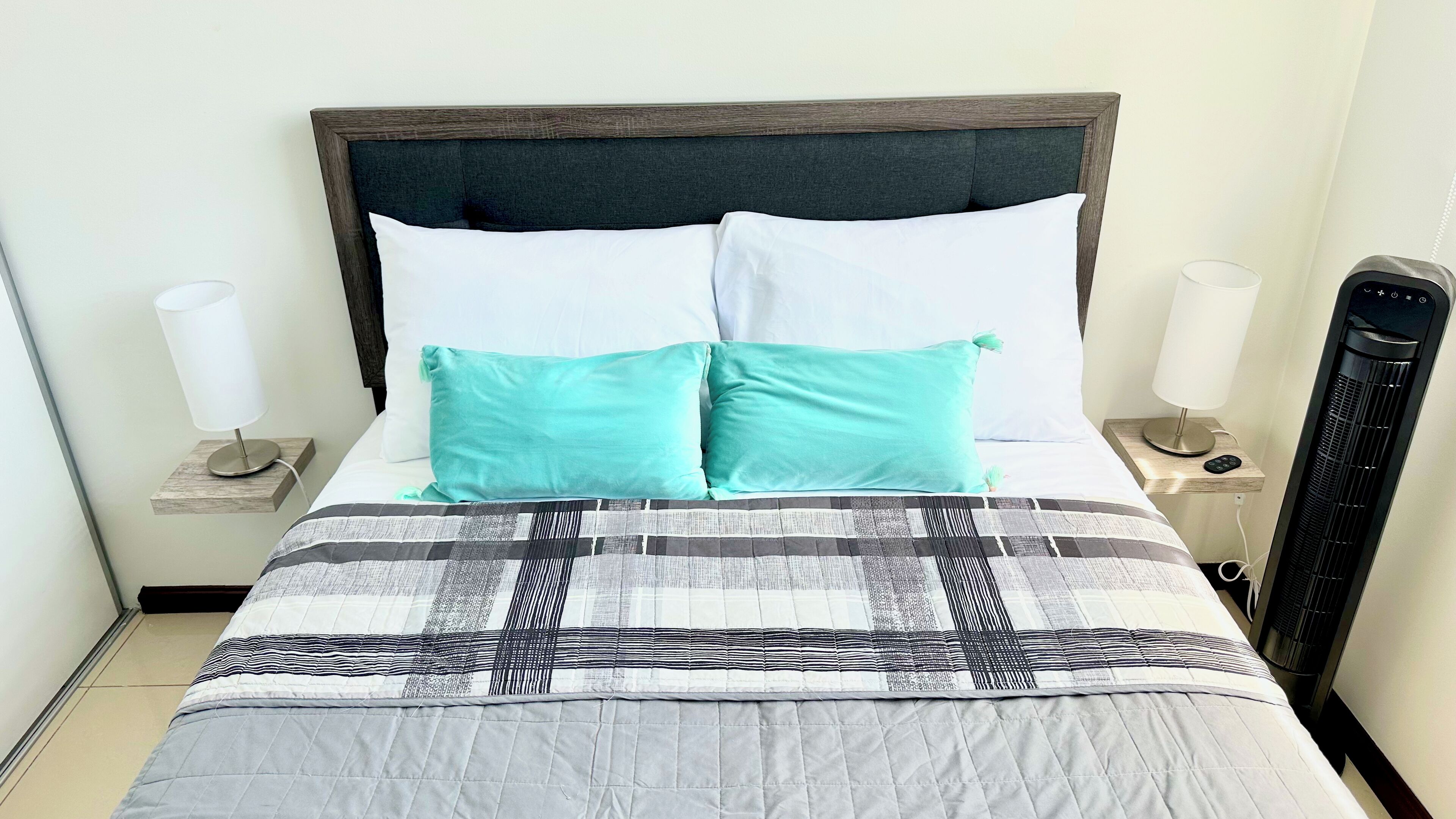 A neatly made bed with a patterned bedspread, two white pillows, and two teal accent pillows. There are bedside tables with lamps and a heater on the right.