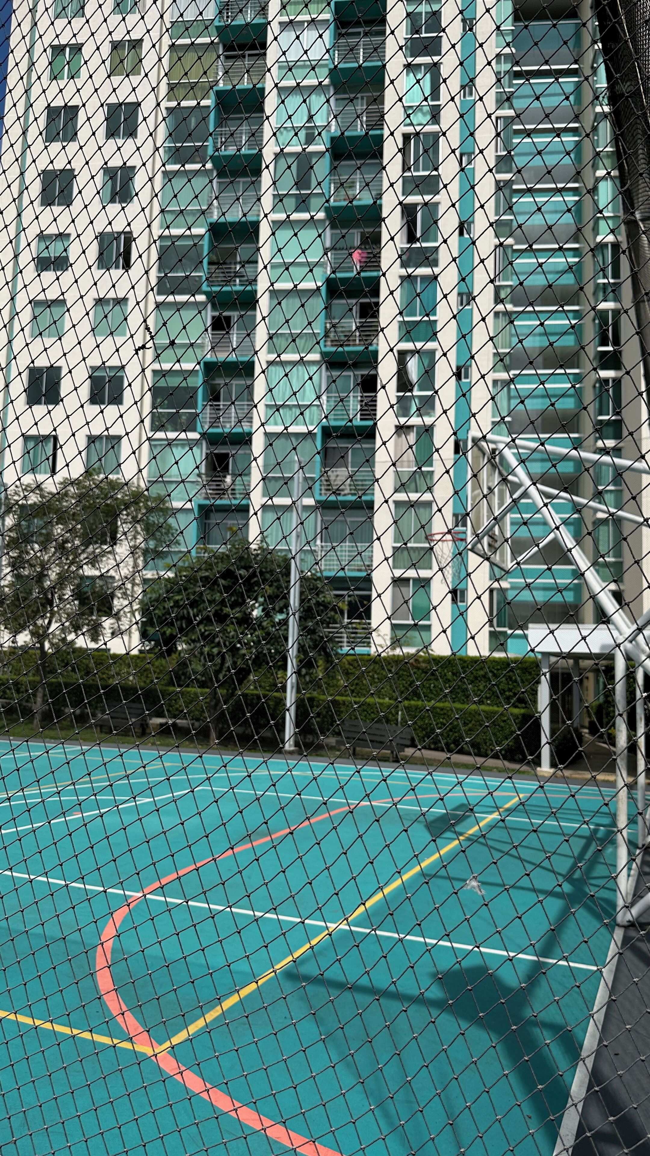 Un terrain de basket entouré d’une clôture en treillis métallique, lui-même bordé par un immeuble de grande hauteur doté de nombreux balcons.