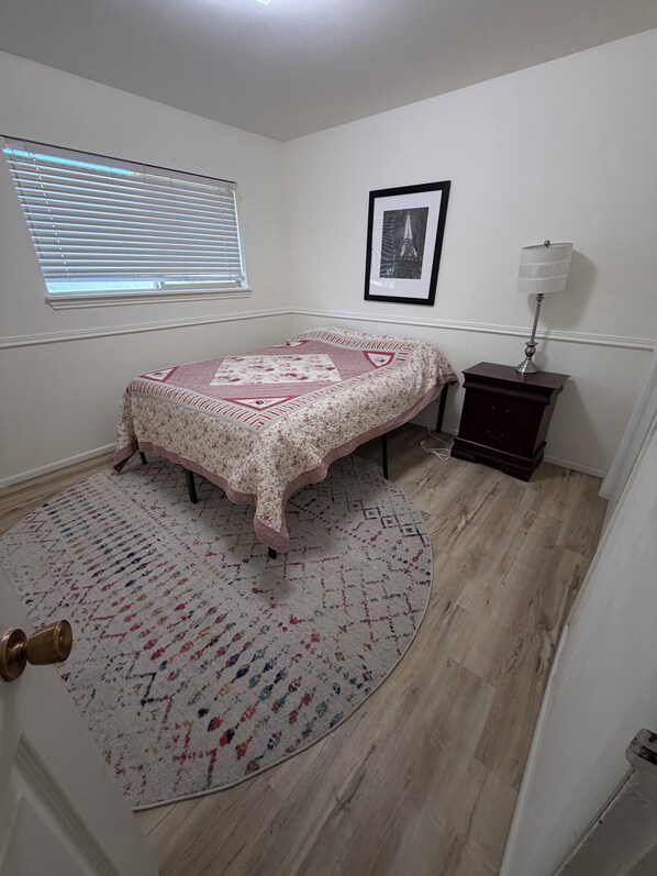 A bedroom with a bed, a bedside table with a lamp, a window with blinds, and a framed picture on the wall.