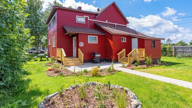 A red two-story house with a front porch, a small attached garage, and a well-maintained garden. - Ely Basecamp- Perfect for Families and Groups (Ely)