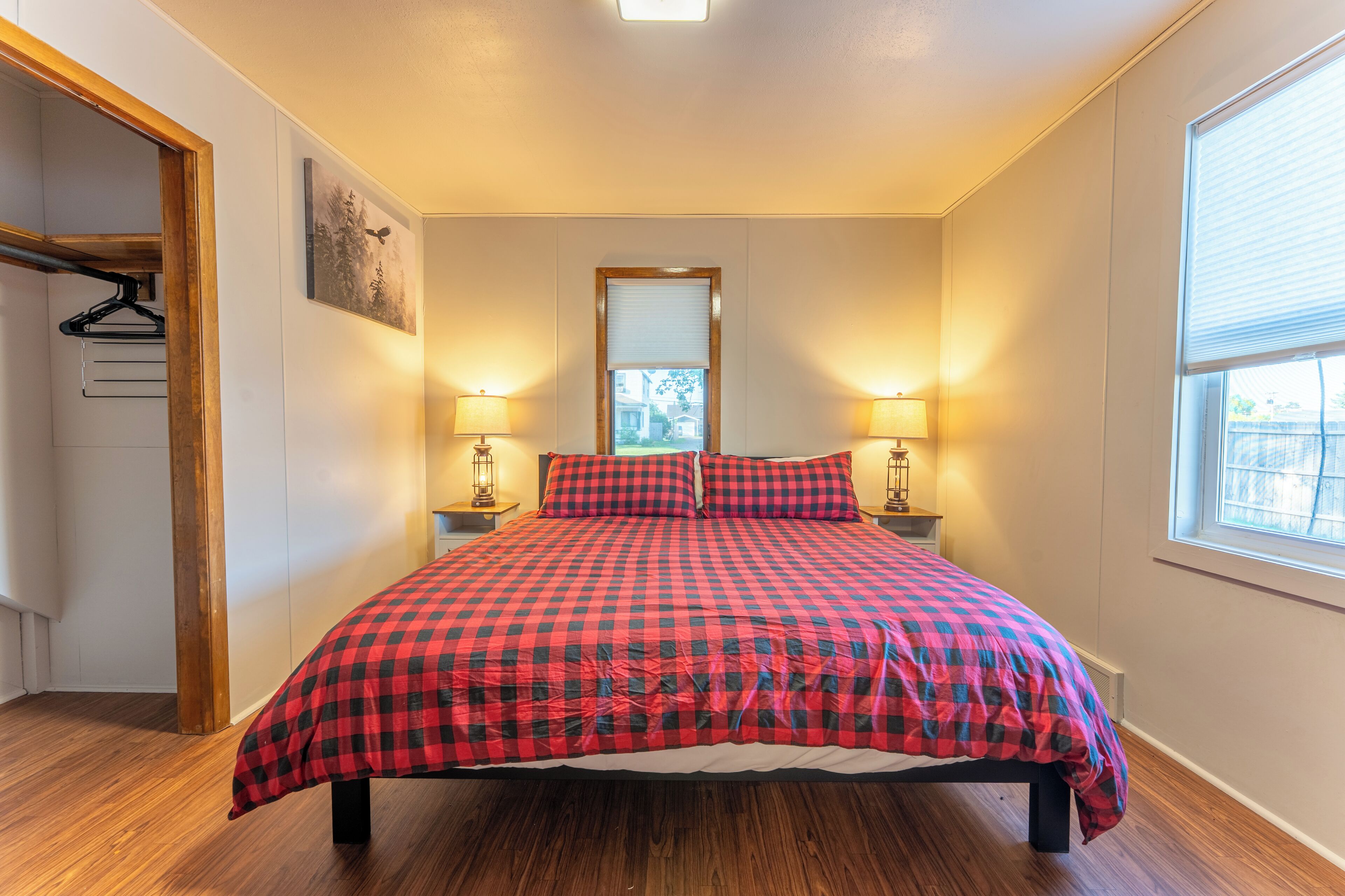 A bedroom with a checkered bedspread, two bedside lamps, a window with blinds, a wooden floor, and a closet with a hanging rack.
