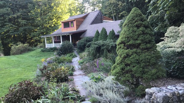 Une maison dotée d’un porche abrité, d’un chemin de pierre et d’une variété de plantes, notamment des conifères et des plantes à fleurs.