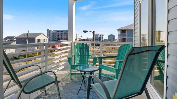 A balcony with green chairs and a table, overlooking a residential area with houses and a street.