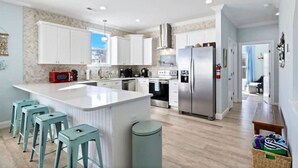 A modern kitchen with white cabinets, a central island with stools, stainless steel appliances, and a view to the living area.