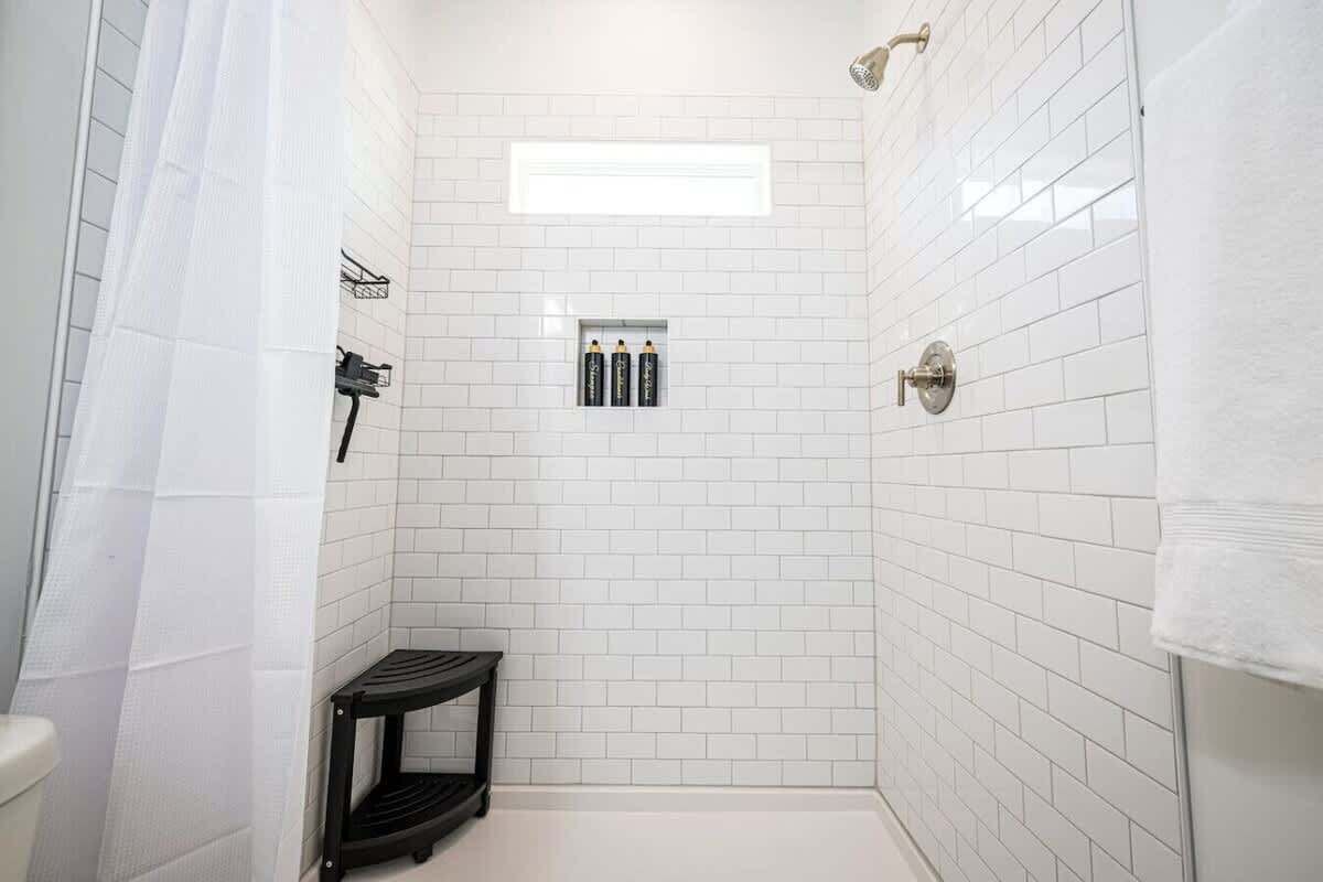 A small, white-tiled shower room with a step stool, a towel rack, and a showerhead.