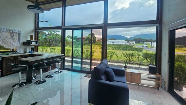 A modern kitchen with a large window, a dining area with a sofa, and a view of a green landscape.