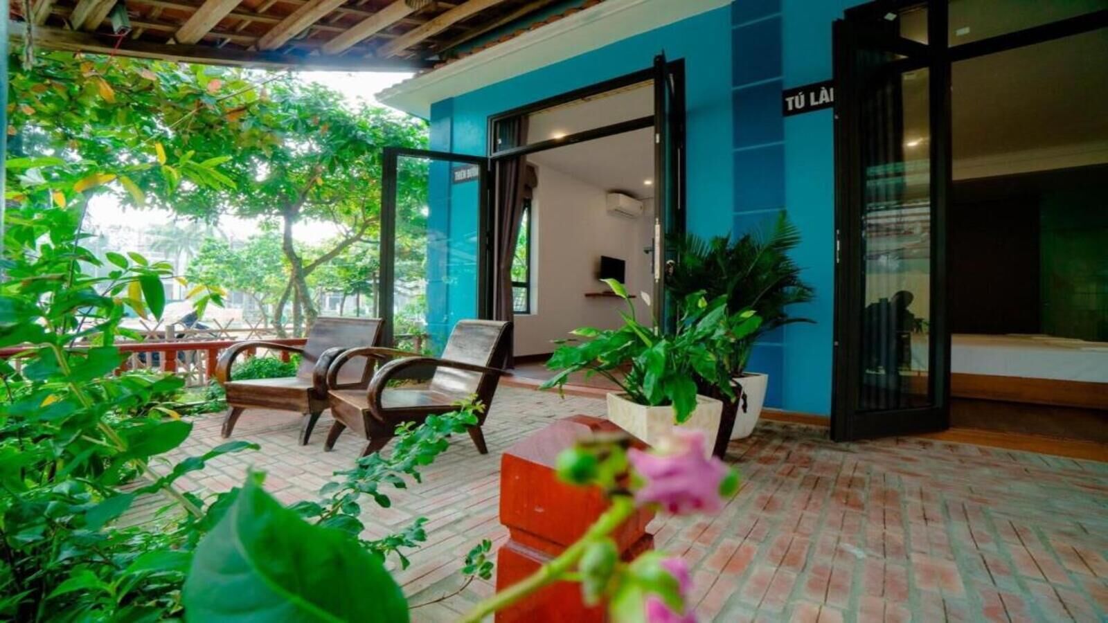 Un espace extérieur aménagé avec des chaises en bois et des plantes en pot, un mur bleu et une enseigne sur laquelle on peut lire « TÜ LÄ ».
