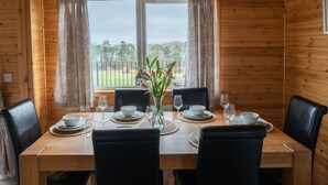 Comedor de una cabaña de madera con una mesa dispuesta para cuatro comensales, dos sillas a cada lado, y vista a un jardín desde una ventana.