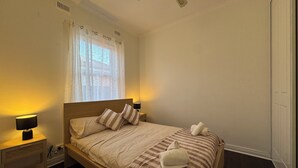 A bedroom with a wooden bed, two bedside lamps, a window with sheer curtains, and a ceiling fan. - Comfy family home(15 mins to CBD & beach) (South Plympton)