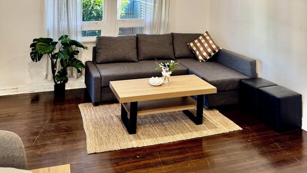 A living room with a sofa, a coffee table, a plant, and a painting on the wall. - Comfy family home(15 mins to CBD & beach) (South Plympton)