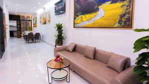 A modern living room with a beige sofa, a glass coffee table with flowers, a dining area in the background, and a large landscape painting on the wall. - CELINA HOTLE Ninh Bình (Hoa Lu)