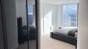 A modern bedroom with a large bed, a bench, and a view of the city through a large window. - Apt Sleeps 6 With Two Bathrooms (Manchester)