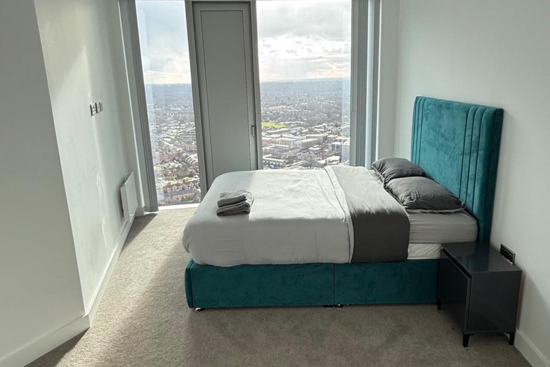 Ein Schlafzimmer mit Bett, Blick auf eine Stadtkulisse durch große Fenster und einem türkisblauen Kopfteil.