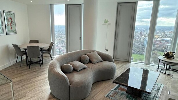 A modern living room with a curved grey sofa, a glass coffee table, and a city view through large windows. - Apt Sleeps 6 With Two Bathrooms (Manchester)