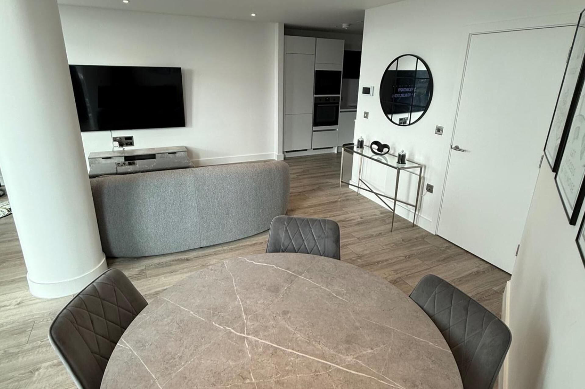 A modern living room with a marble dining table, grey cushioned seating, a flat-screen TV mounted on the wall, and a kitchen area with white cabinets and a round mirror.