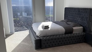 A bedroom with a large window, a bed with a dark headboard, and a view of a cityscape. - Apt Sleeps 6 With Two Bathrooms (Manchester)