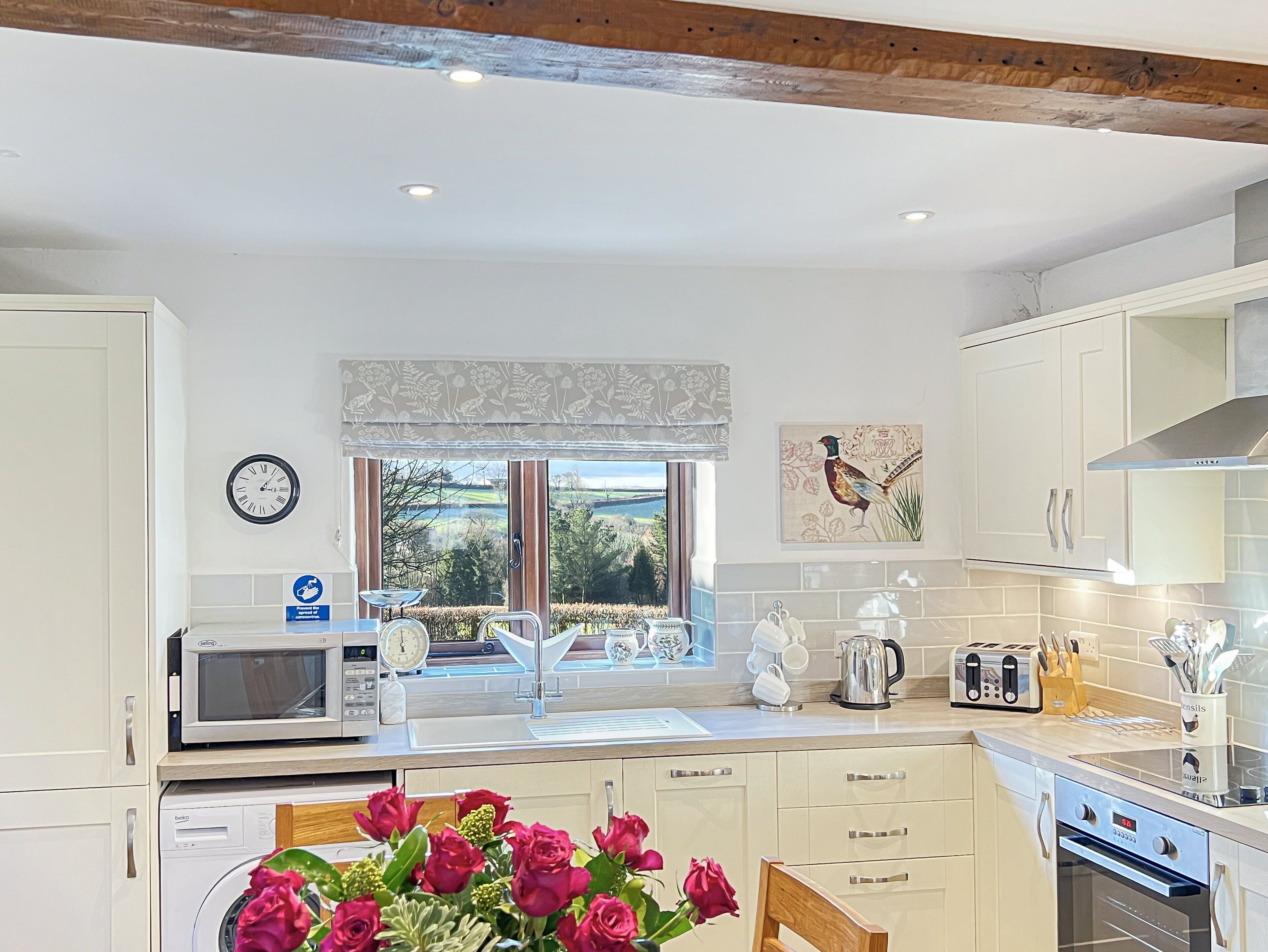 A modern kitchen with a built-in oven, microwave, and sink. There is a window with a view of the countryside, and a clock on the wall.