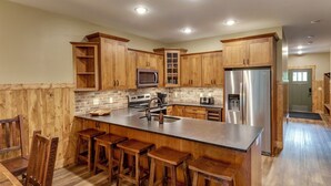 A kitchen with wooden cabinets, a stainless steel refrigerator, a microwave, and a sink. There are wooden stools around the island.