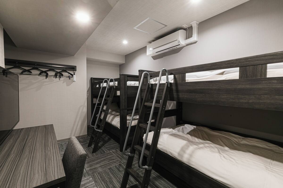 A dormitory room with bunk beds, a desk, and a chair. There is a wall-mounted air conditioner and a light fixture.
