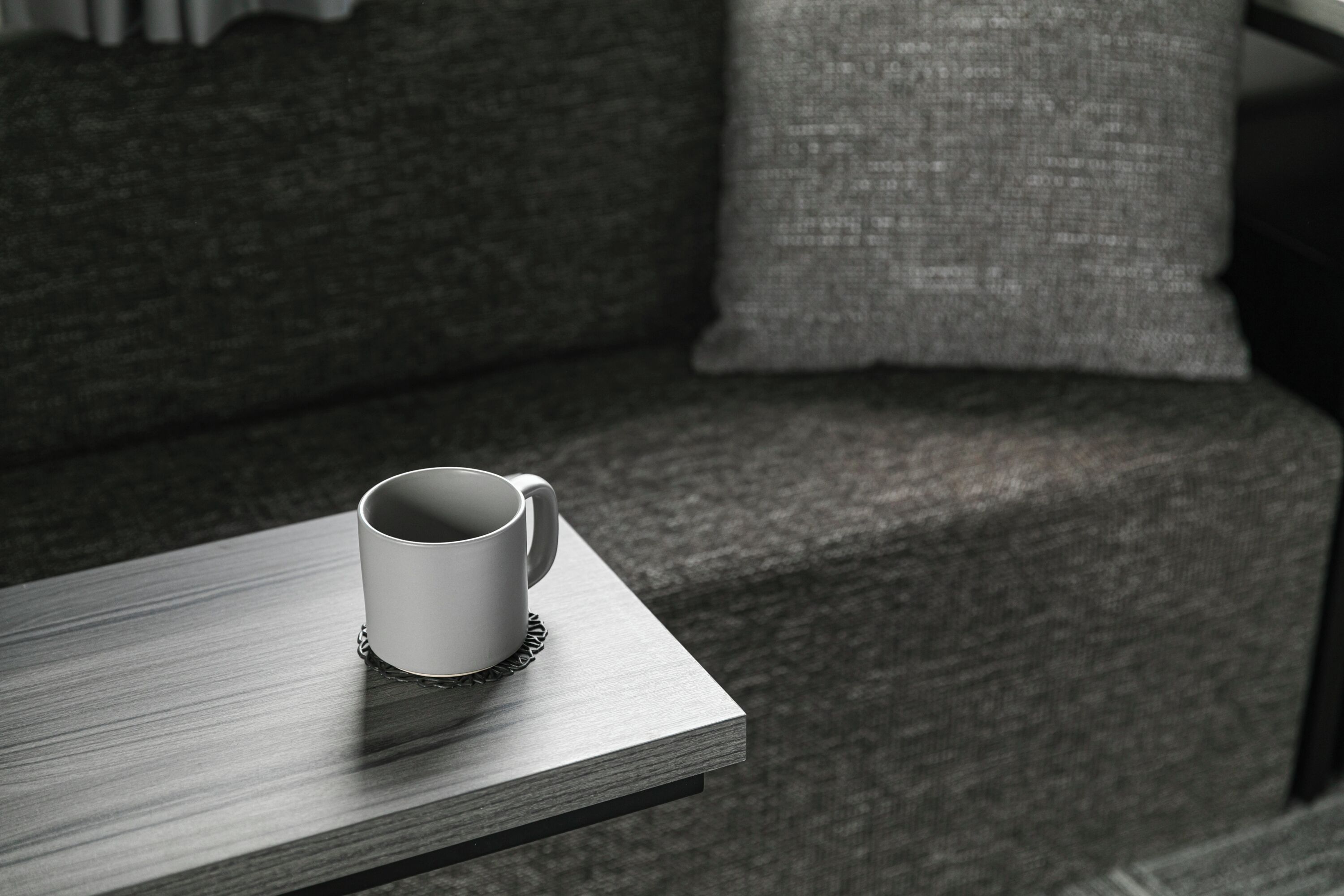 Een witte mok op een houten tafel in een kamer met een grijze bank en een grijs kussen.