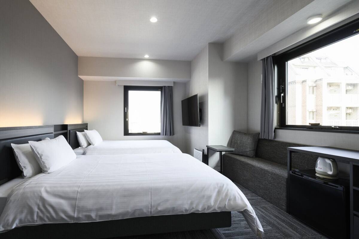 A hotel room with a large bed, a TV, a seating area, and a window with a view of buildings.