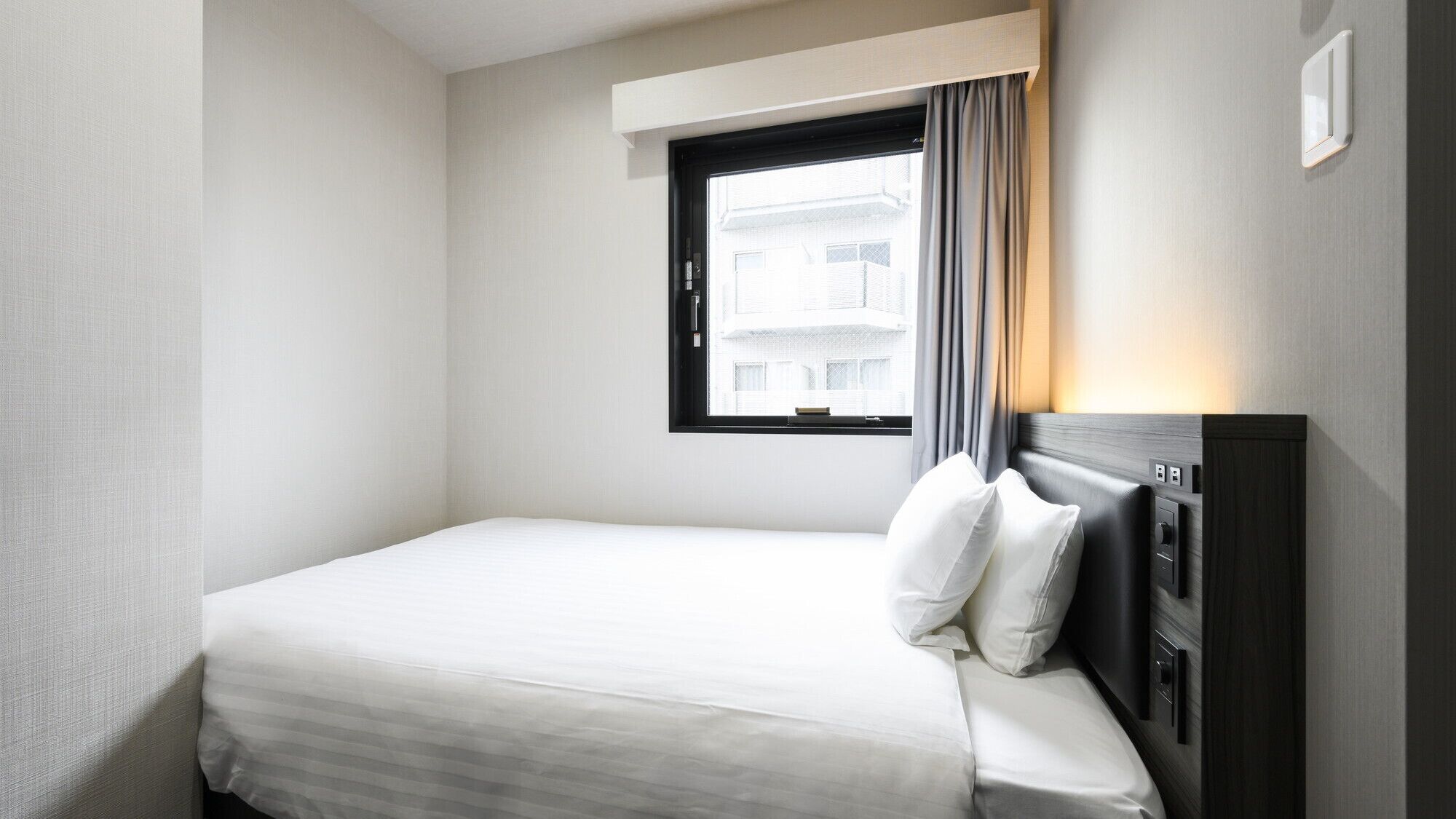 Une chambre d’hôtel avec un lit simple, une fenêtre avec des rideaux, une table de chevet et une lampe fixée au mur.
