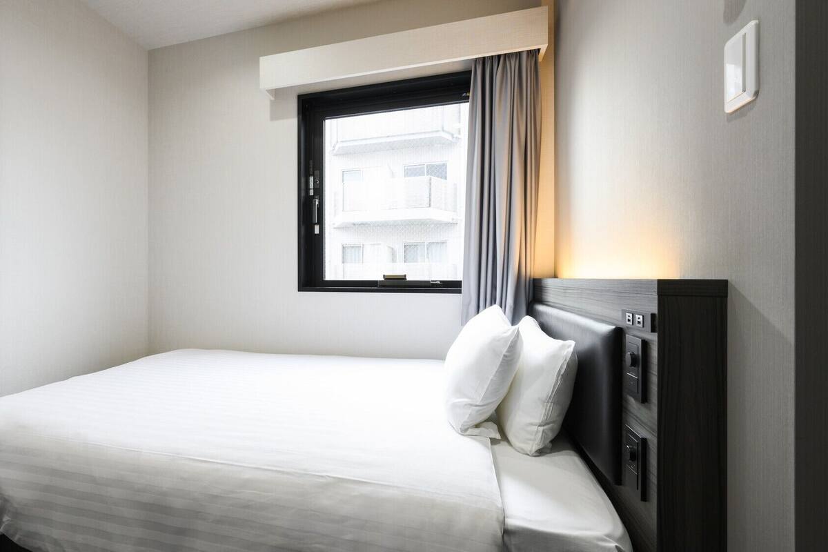 A hotel room with a single bed, a window with curtains, a bedside table, and a wall-mounted light.