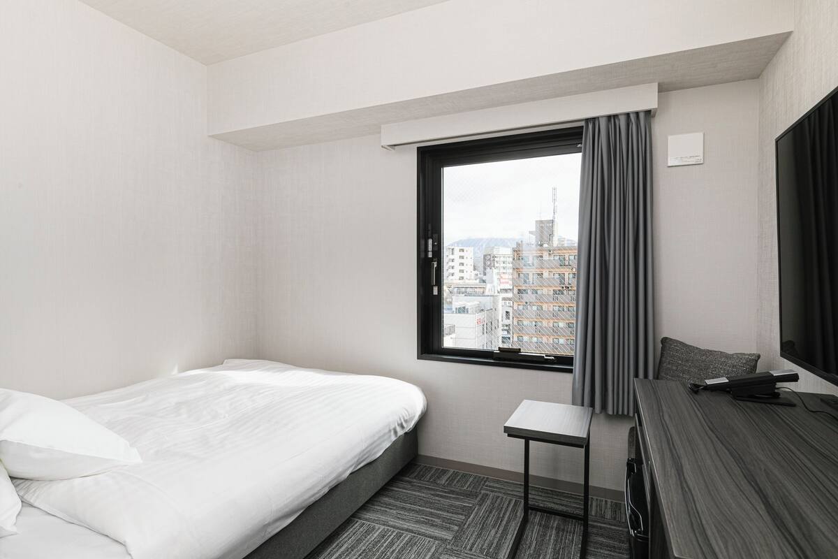 A hotel room with a bed, a small table, a desk, a window with curtains, and a view of buildings.