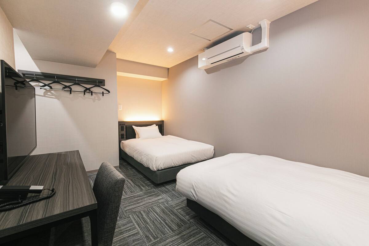 A hotel room with a desk, two beds, a chair, and an air conditioning unit.