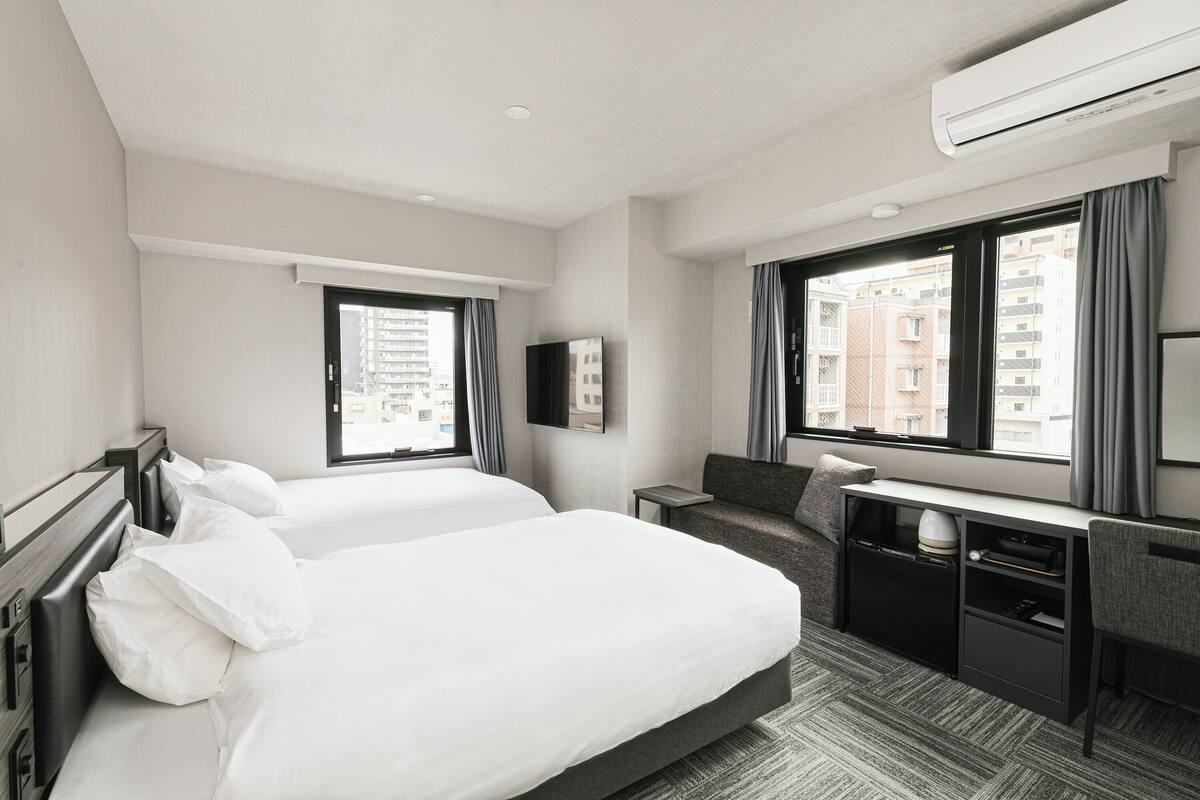 A hotel room with two beds, a desk, a chair, a flat-screen TV, and large windows with curtains.
