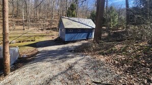 Une petite cabane à un seul étage, avec un toit en métal, entourée d’arbres et bordée de chemins de gravier.