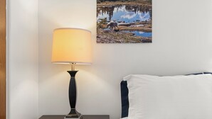 Una cama bien hecha con ropa de cama blanca y azul oscuro, una lámpara de noche en una mesita de madera, y una fotografía en el marco de un paisaje natural en la pared.