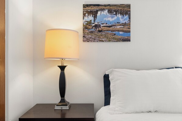 A neatly made bed with white and dark blue bedding, a bedside lamp on a wooden nightstand, and a framed photograph of a natural landscape on the wall. - LoHi Home – Walk to Dining, Bars, Downtown and more! (Denver)