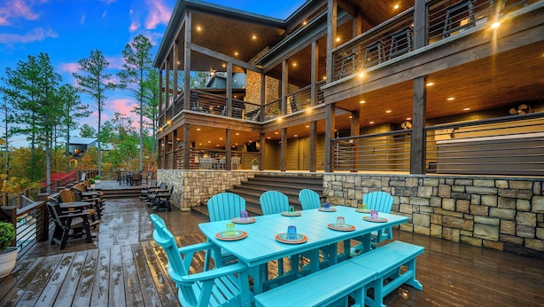 A wooden deck with a dining table and chairs, surrounded by stone walls and a wooden railing. - Inspiration Point | 6 King Suites, 2 Bunk Rooms, Pool, 2 Hot Tubs, Pickleball & Creek Views (Broken Bow)