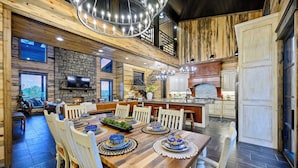 A spacious dining area with a wooden table set for a meal, a kitchen with a stone backsplash, and a large chandelier. - Inspiration Point | 6 King Suites, 2 Bunk Rooms, Pool, 2 Hot Tubs, Pickleball & Creek Views (Broken Bow)