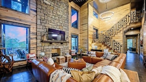 A spacious living room with a stone wall, leather sofas, a TV, and a staircase. - Inspiration Point | 6 King Suites, 2 Bunk Rooms, Pool, 2 Hot Tubs, Pickleball & Creek Views (Broken Bow)
