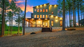 A modern two-story house with a stone foundation, multiple windows, and a porch with outdoor seating. - Luxury Cabin | Heated Pool, Hot Tub, Game Loft, Pickleball Court | Sleeps 23 – Risky Business (Broken Bow)