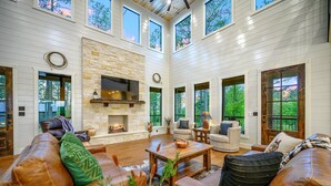 A spacious living room with a stone fireplace, leather sofas, and large windows offering views of the outdoors. - Luxury Cabin | Heated Pool, Hot Tub, Game Loft, Pickleball Court | Sleeps 23 – Risky Business (Broken Bow)