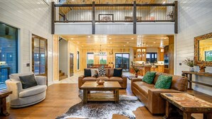 A spacious living area with a high ceiling, wooden flooring, and a large window providing a view of the outdoors. - Luxury Cabin | Heated Pool, Hot Tub, Game Loft, Pickleball Court | Sleeps 23 – Risky Business (Broken Bow)