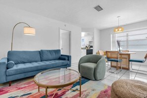 Ein modernes Wohnzimmer mit einem blauen Sofa, einem runden Glastisch, einem grauen Sessel und einem Essbereich mit Korbstühlen und einem Tisch.