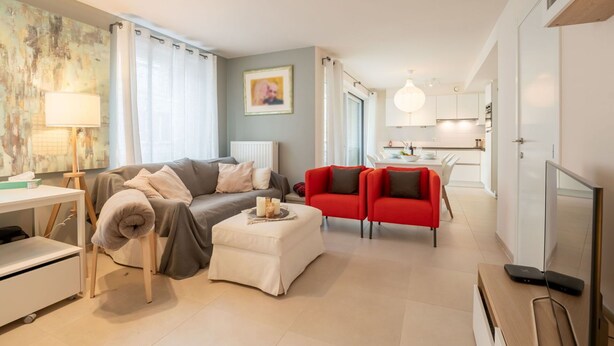 Una sala de estar moderna con un sofá, un reposapiés y sillas rojas. Hay un comedor con una mesa y sillas, y una cocina al fondo.