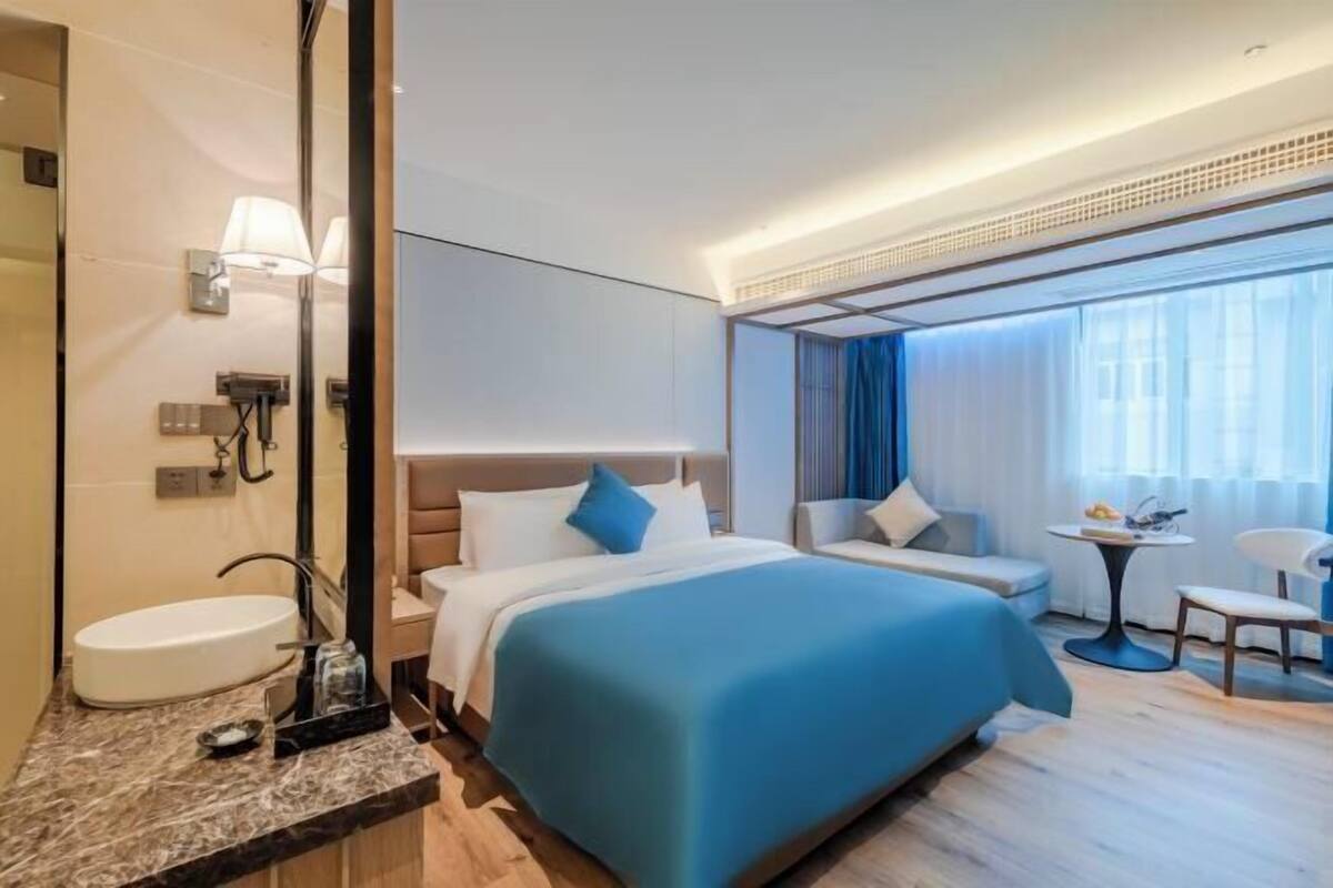 Une chambre d’hôtel dotée d’un grand lit, d’un coin salon avec une table et des chaises, d’une salle de bain avec un lavabo et un miroir, et d’une grande fenêtre avec des rideaux.