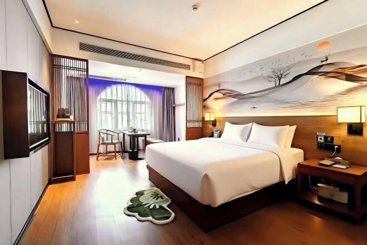 Une chambre d’hôtel avec un grand lit, un bureau et une chaise. Il y a un tableau au mur et une fenêtre avec des rideaux.