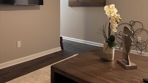 Una sala de estar con un televisor de pantalla plana montado en la pared, una mesa de centro de madera con objetos decorativos, y un cuadro de un árbol.