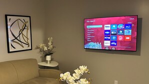 Una sala de estar con un televisor de pantalla plana montado en la pared mostrando la interfaz de un servicio de transmisión, un sofá color beige, una mesita auxiliar con una orquídea en maceta y una obra de arte abstracta enmarcada.