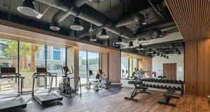 A modern gym with various exercise equipment, large windows, and a high ceiling.