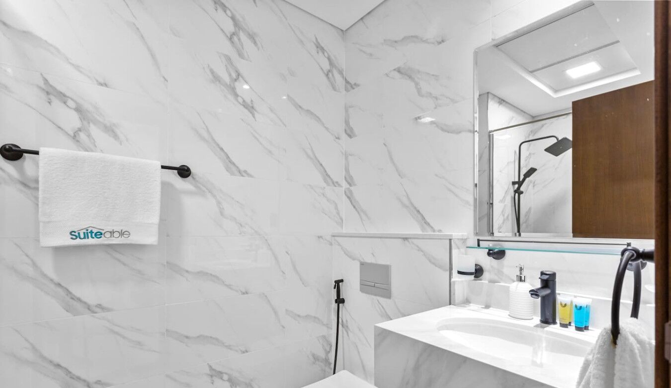 A modern bathroom with a marble wall, a glass shower enclosure, a white sink with toiletries, and a towel hanging on a rack.