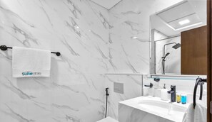 A modern bathroom with marble walls, a glass shower enclosure, a white sink with toiletries, and a towel rack with a 'Suiteable' branded towel.