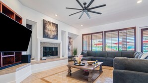 A living room with a flat-screen TV, a fireplace, a ceiling fan, and a large window with blinds. - ALL INCLUSIVE - SUPER MARIO RETREAT – EPIC GAMING GETAWAY (Indio)