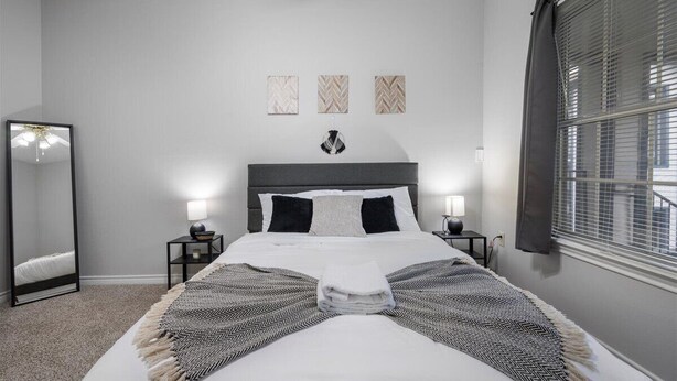 Un dormitorio ordenado con una cama grande, dos mesitas de noche con lámparas, un espejo y tres obras de arte colgadas en la pared.